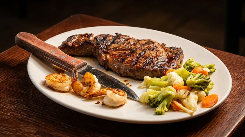 Juicy grilled steak, seasoned shrimp, and steamed vegetables on a white plate with a knife.